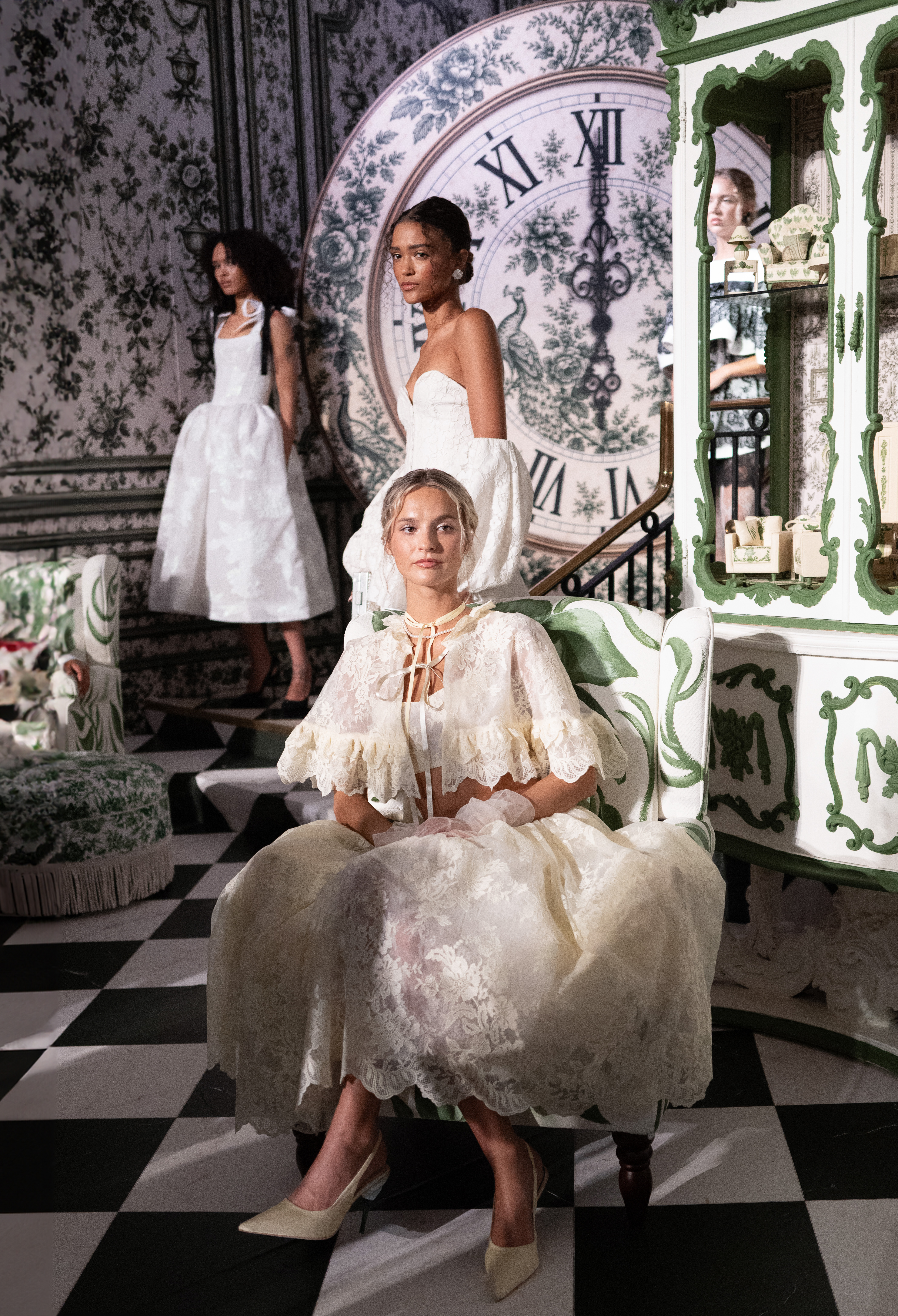 Three models in white lace, brocade, and tulle gowns pose in front of an oversized painted clock at Alice + Olivia's Wonderland presentation.