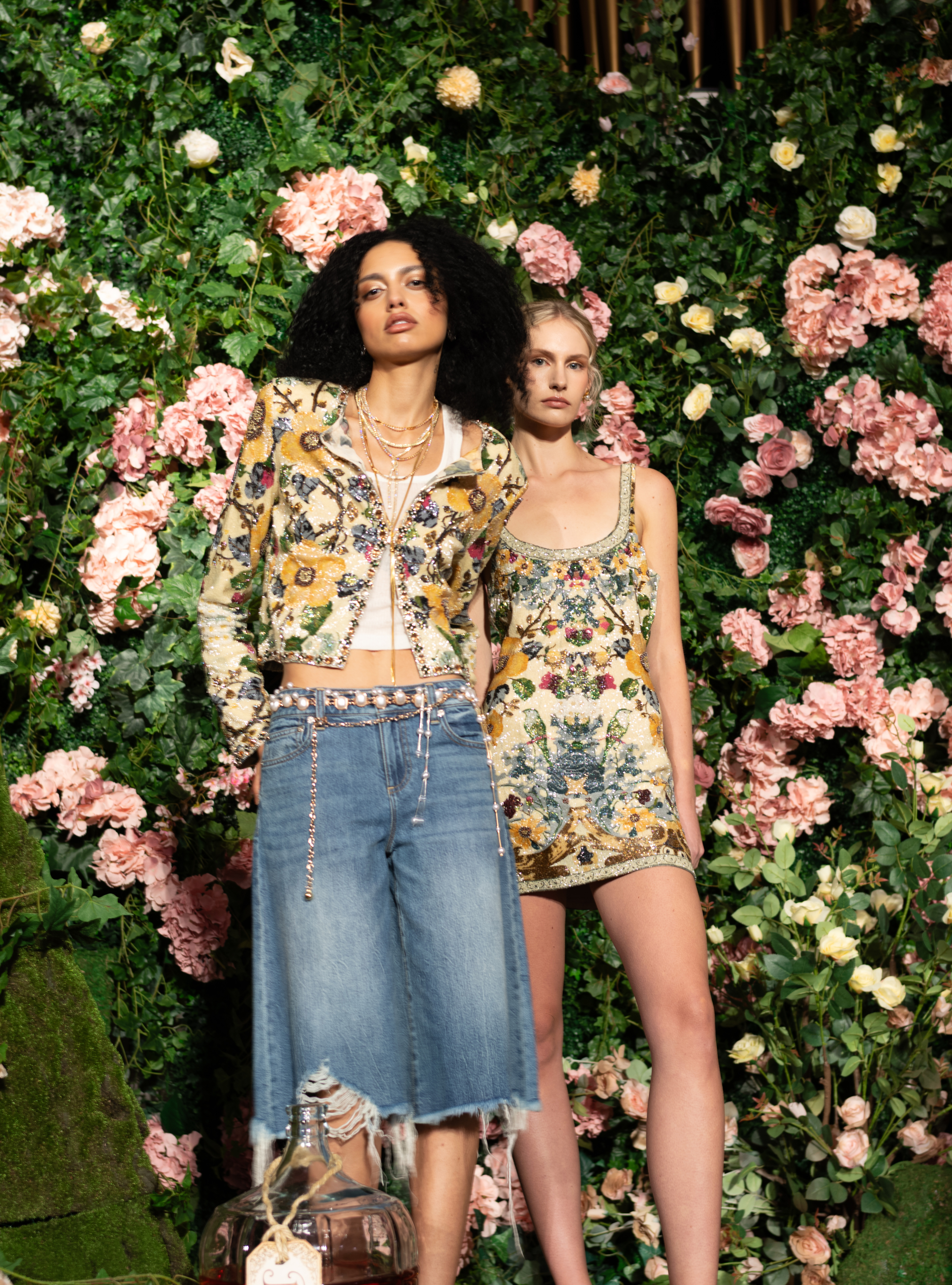 Two looks from Alice + Olivia's Wonderland collection pairing beaded floral embroidery with wide-leg denim and a matching sequined mini dress, set against a wall of roses.