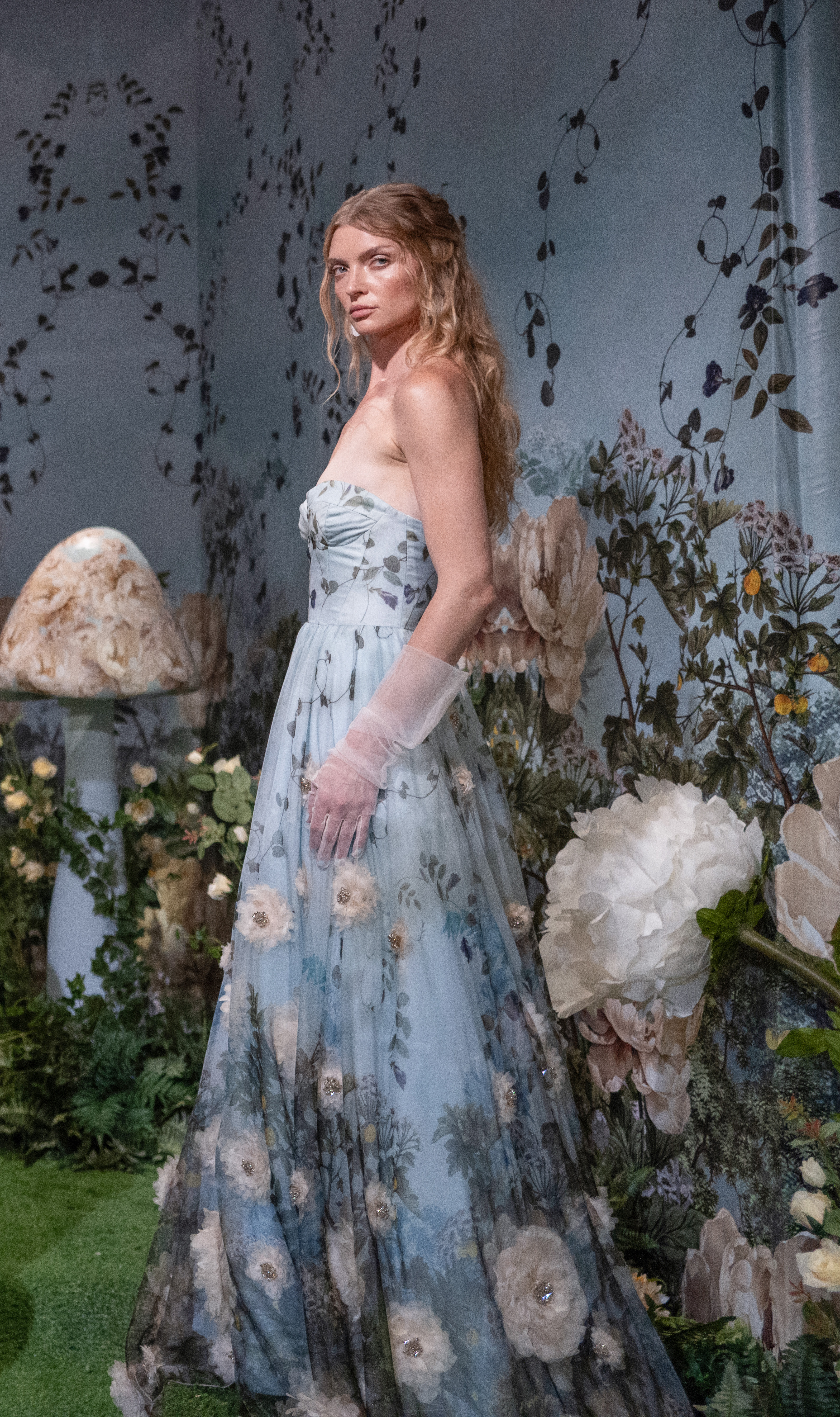 A strapless, floral-embellished tulle gown in pale blue from Alice + Olivia's Wonderland collection, styled with sheer opera gloves against a garden backdrop.
