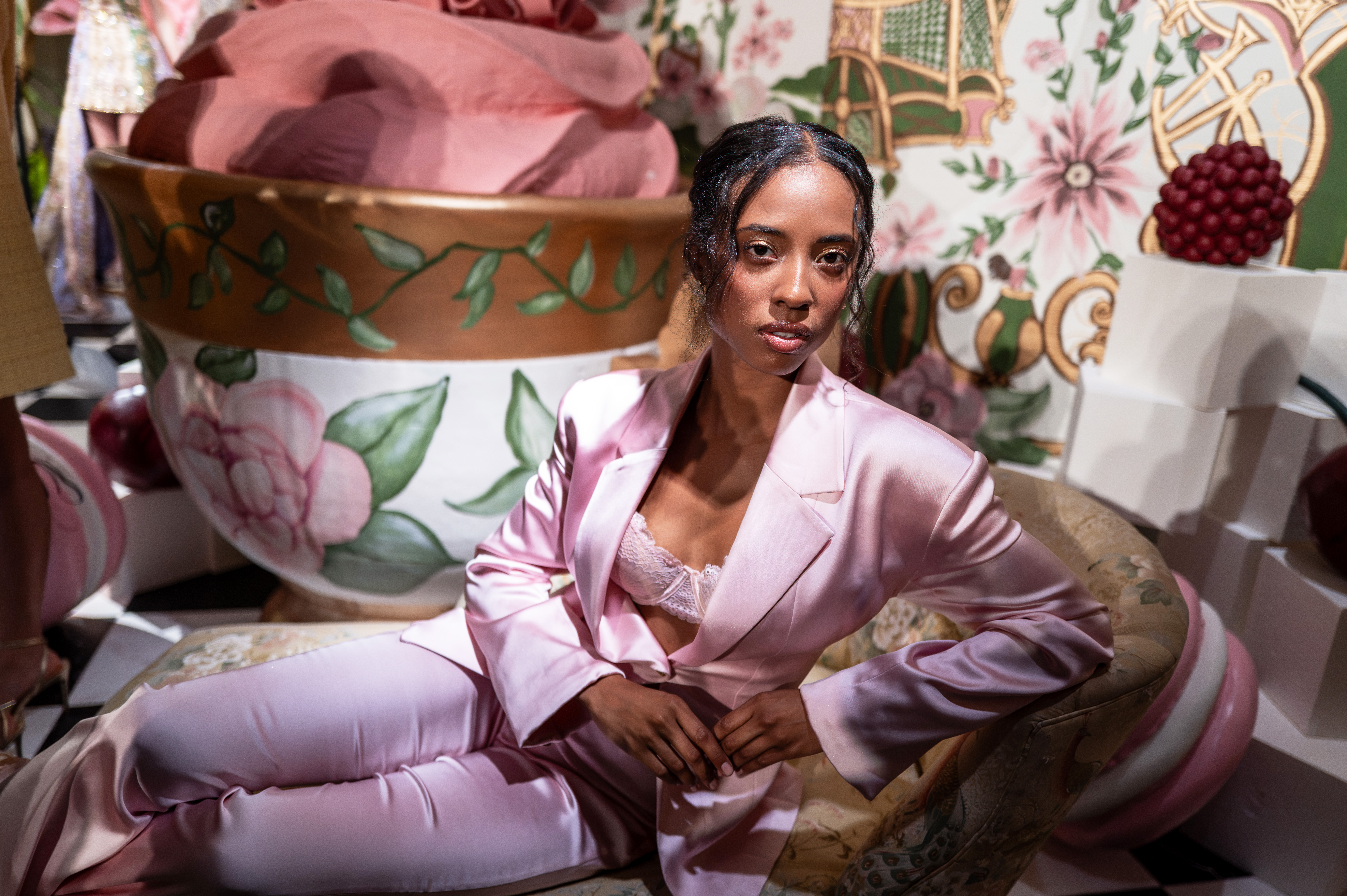 A model in a pale pink satin pajama-style suit reclines beside an oversized hand-painted teacup prop at Alice + Olivia's Wonderland presentation.