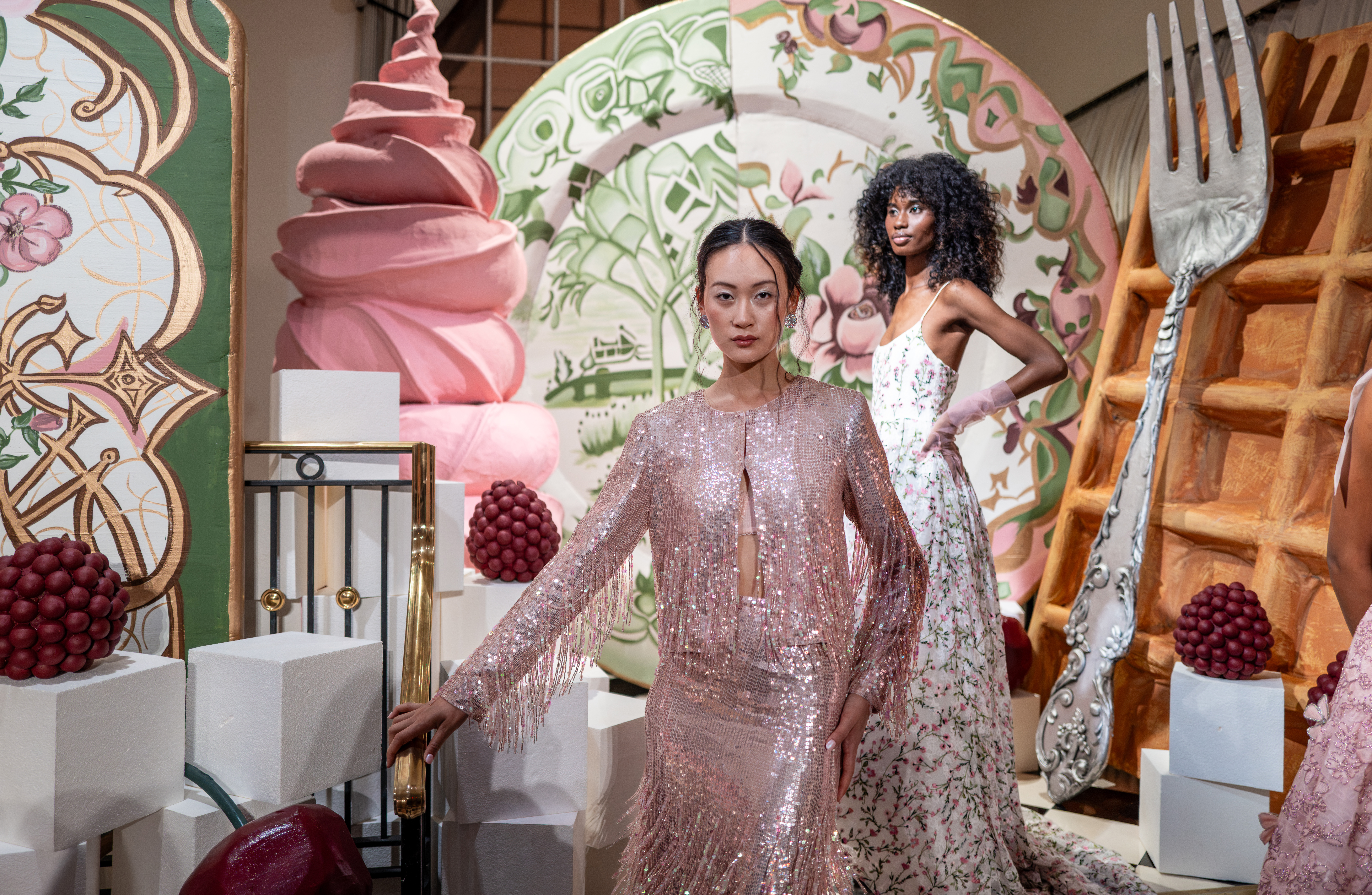 Two models in a sequined pink fringe gown and a floral spaghetti-strap dress pose beside an oversized soft-serve sculpture and giant silver fork at Alice + Olivia's Wonderland presentation.