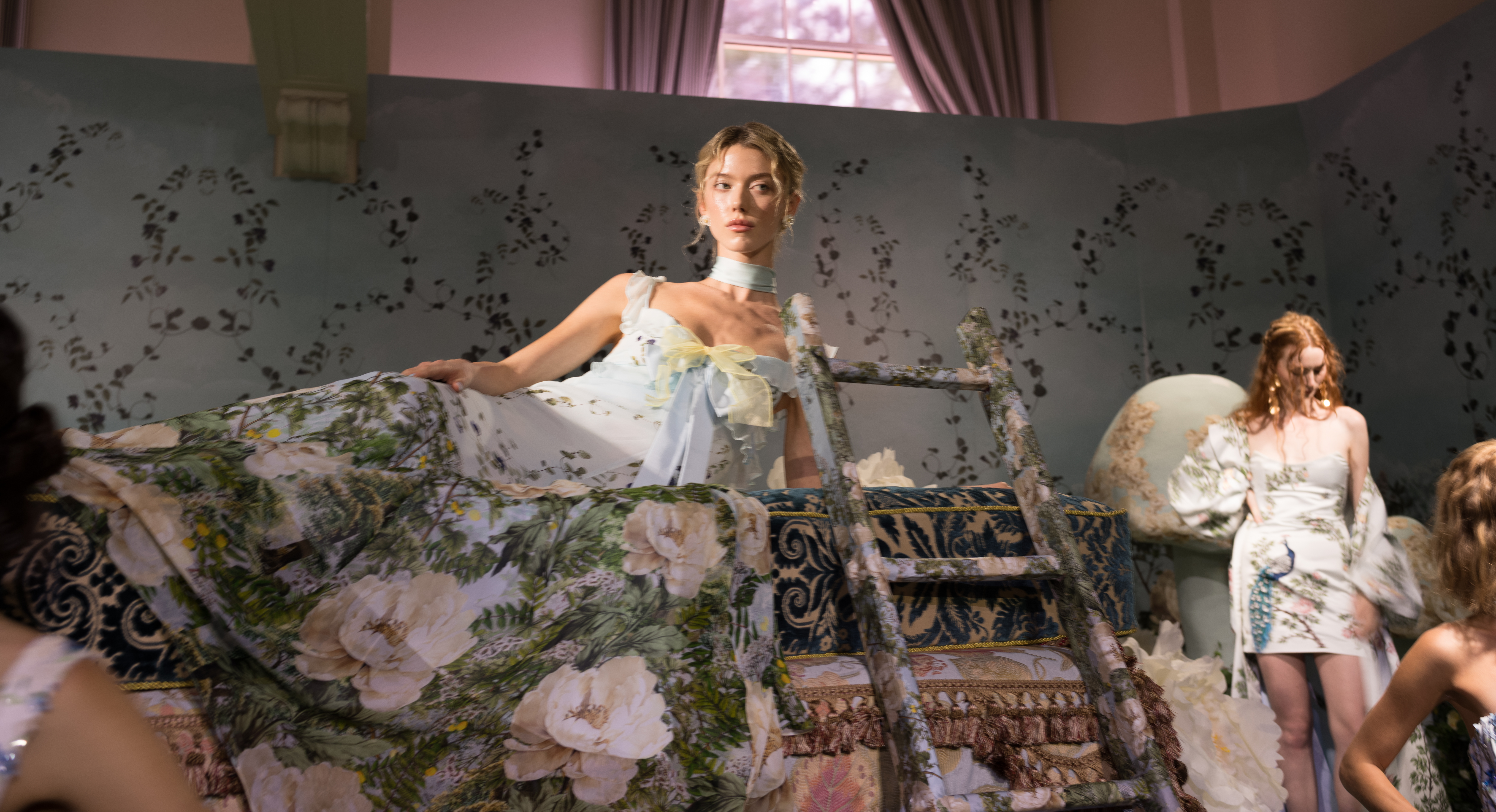 A model in a pale blue bow-detailed gown reclines on an antique floral settee backstage at Alice + Olivia's Wonderland presentation.