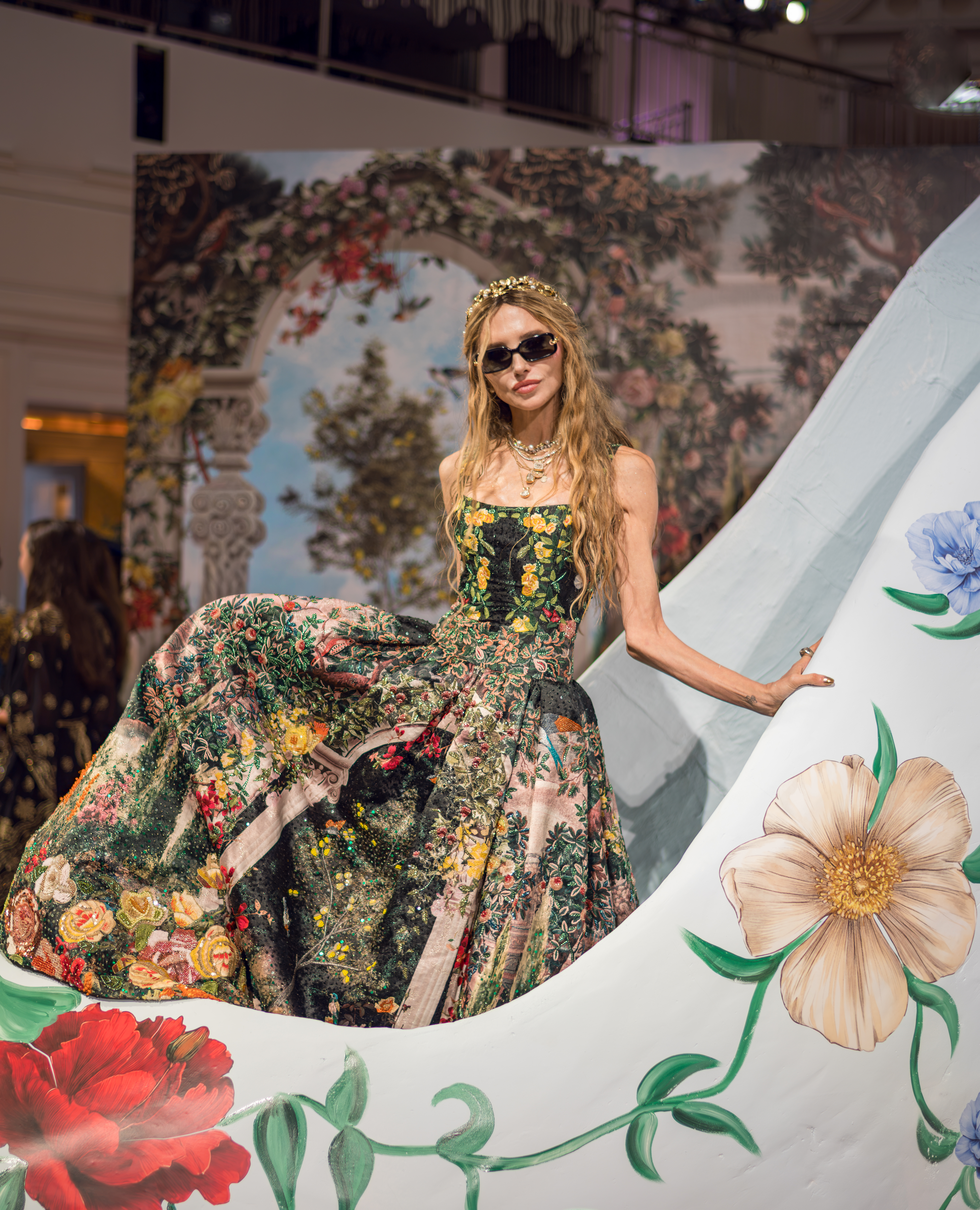 A model in a maximalist dark floral ball gown, sunglasses, and a gold laurel headpiece at Alice + Olivia's Wonderland presentation.