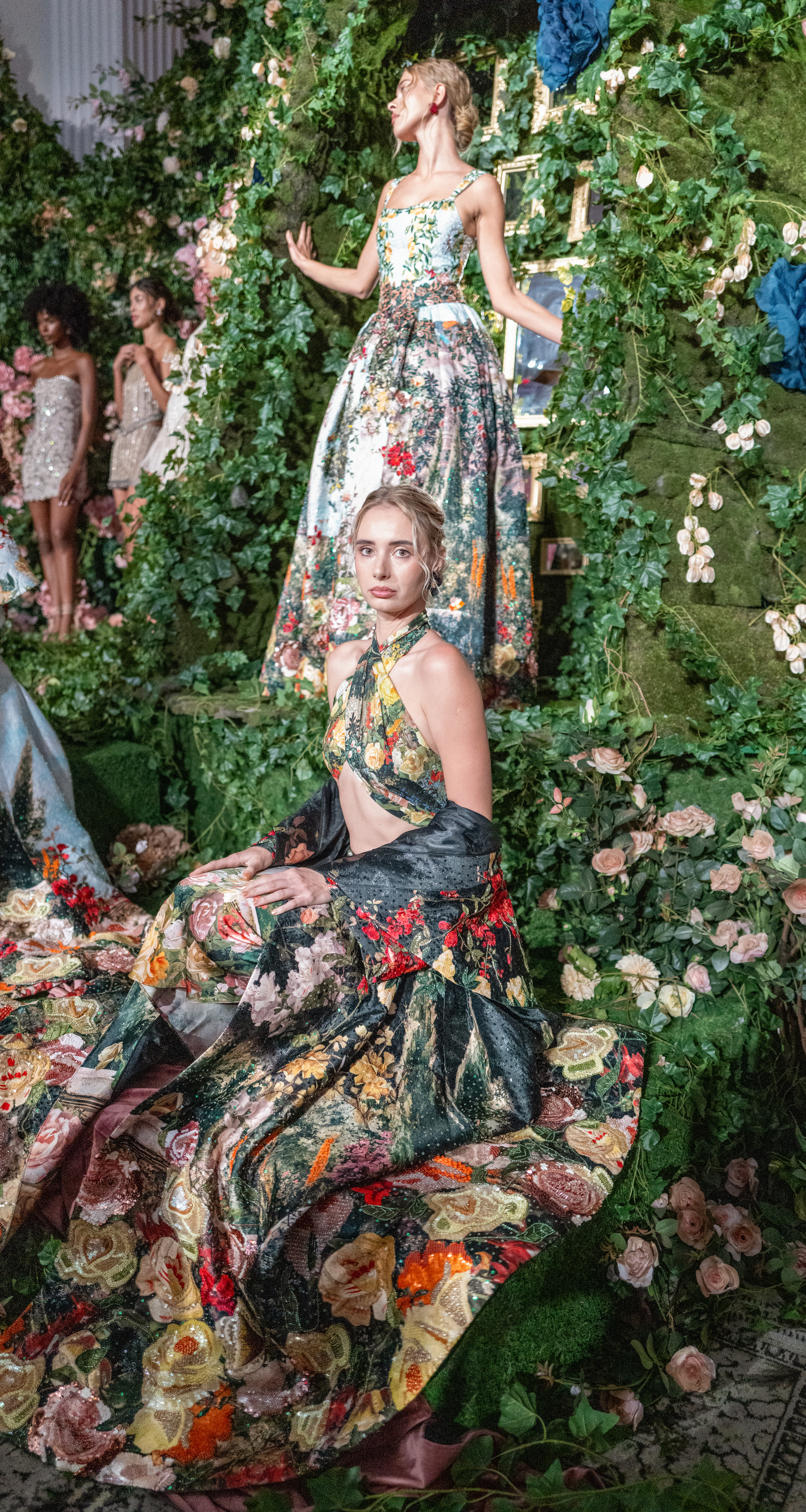 Two models in floral ball gowns — one ivory, one black-floral with a halter neckline — on a moss-and-rose garden set at Alice + Olivia's Wonderland presentation.